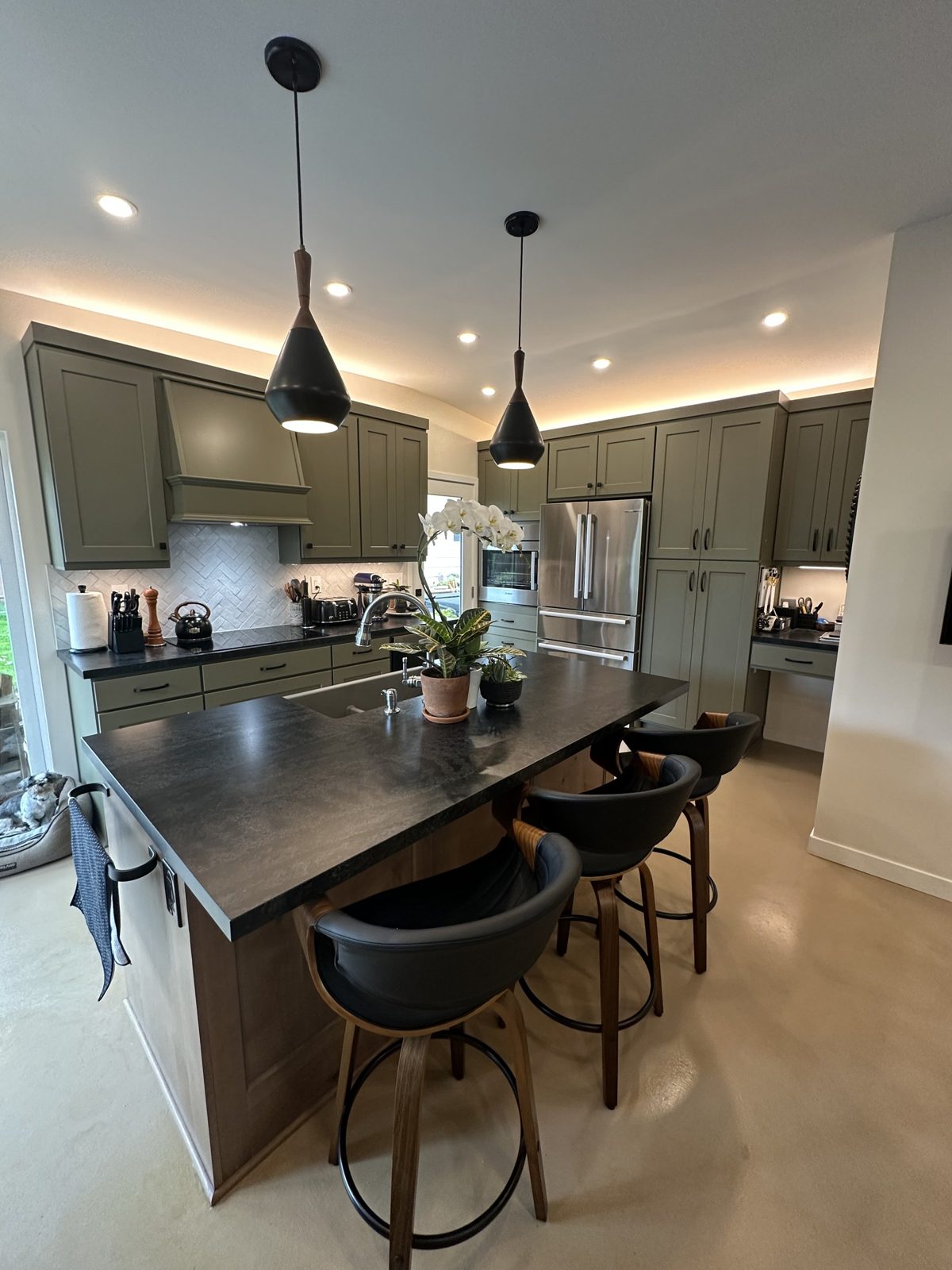 New kitchen island with honed black countertop, swivel bar stools and sage green Shaker cabinets with cove lighting