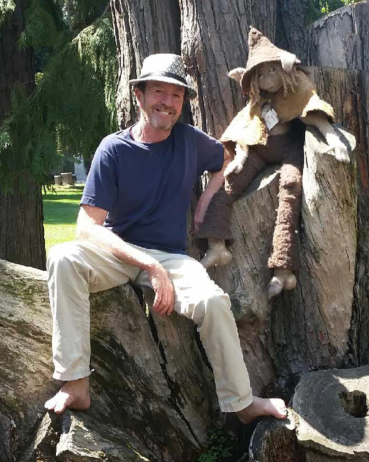 Jonathan Quintin sitting barefoot on a large tree stump outdoors, with a small felted gnome beside him
