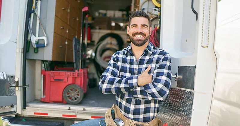Home-services contractor standing at the open rear doors of his stocked work van, arms crossed and smiling