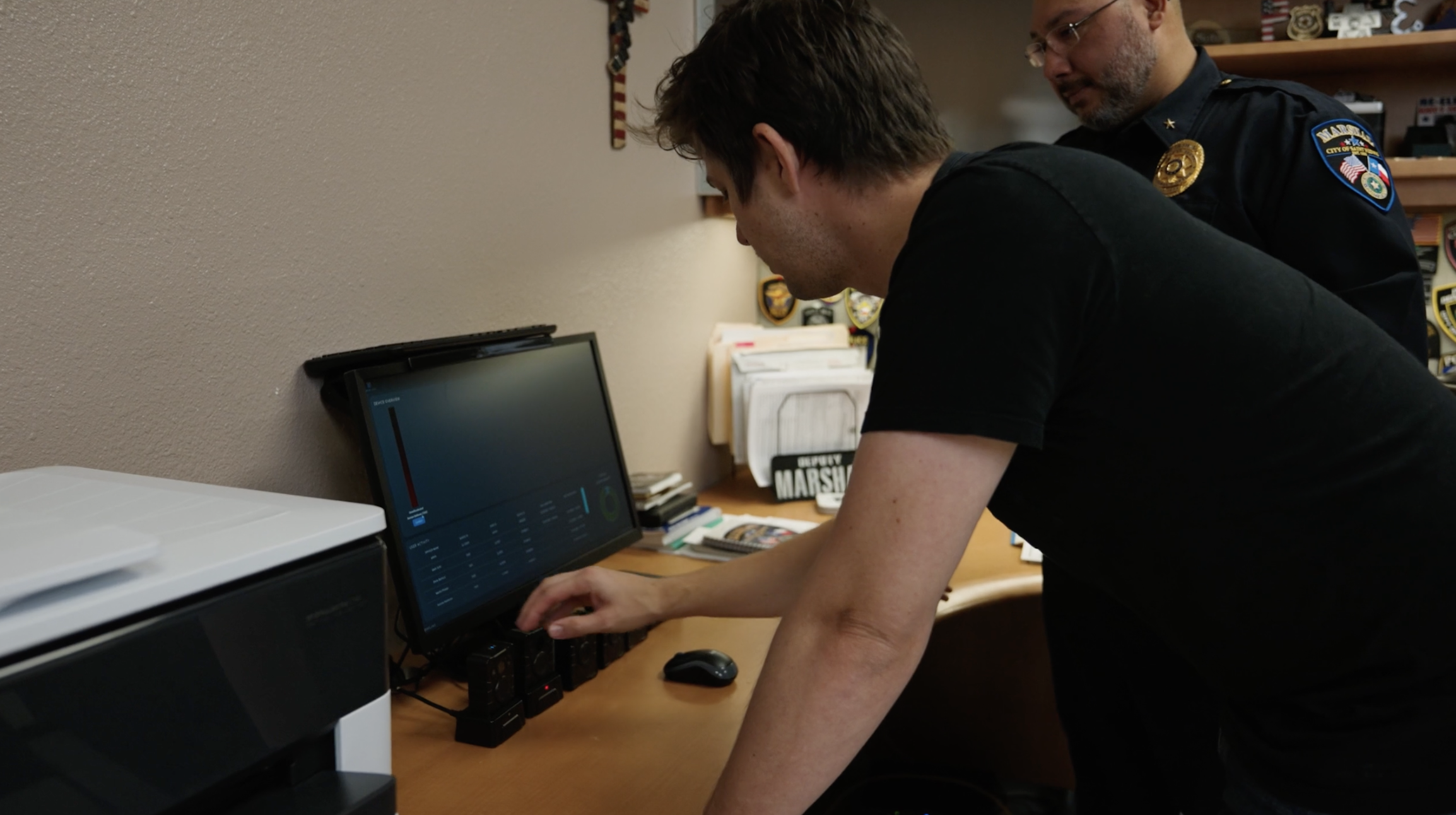 Technician placing body cameras in a docking station at a marshal's office desk while a uniformed marshal looks on