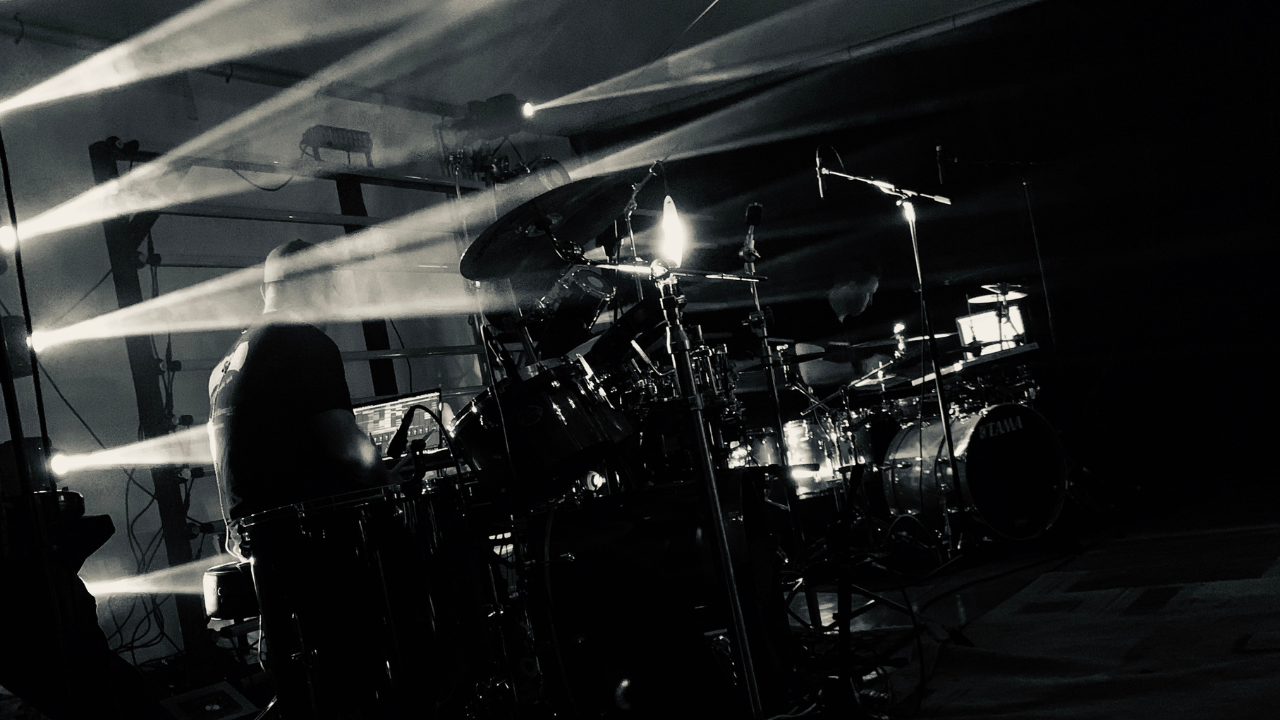 Tim Alexander performing behind his drum kit under dramatic stage lighting. Tim Alexander performing behind his drum kit under dramatic stage lighting.