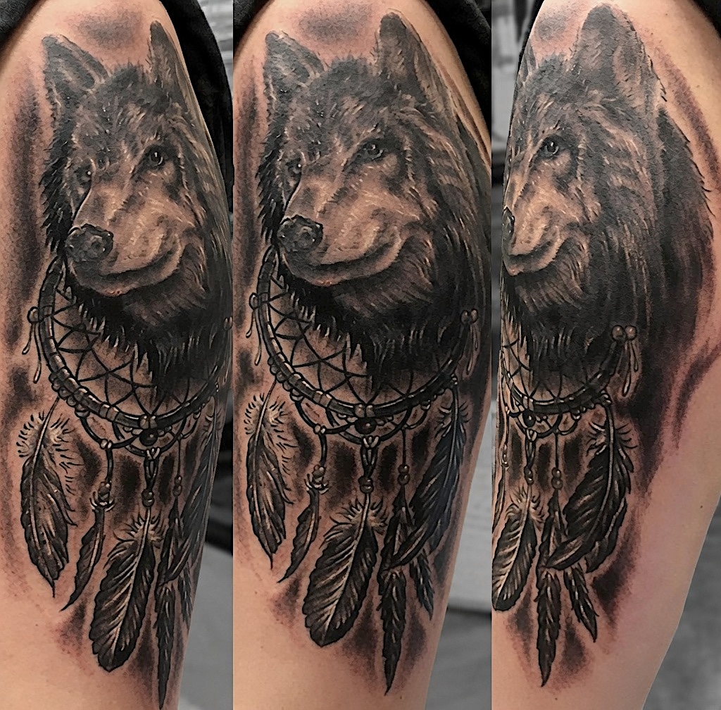 Black and grey wolf portrait tattoo with a feathered dreamcatcher on the upper arm