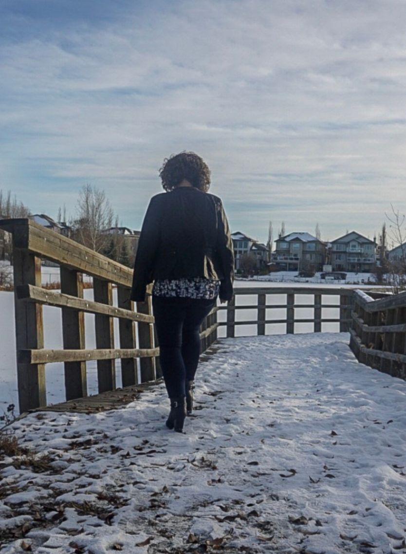 Leanne Chesser wearing blue jeans, a black leather jacket and a shirt with blue and maroon irregular shapes. She's walking away from the camera down a snow-covered pathway with a wooden railing. There are houses in the background.