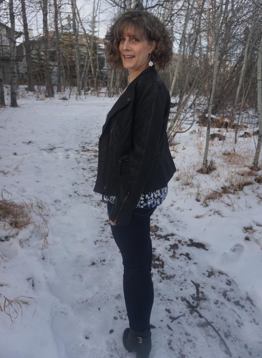 Leanne Chesser wearing blue jeans, a black leather jacket, black leather boots and a shirt with blue and maroon irregular shapes. She's walking away from the camera down a snow-covered path and turning back to look over her left shoulder. There are bare trees and houses in the background.