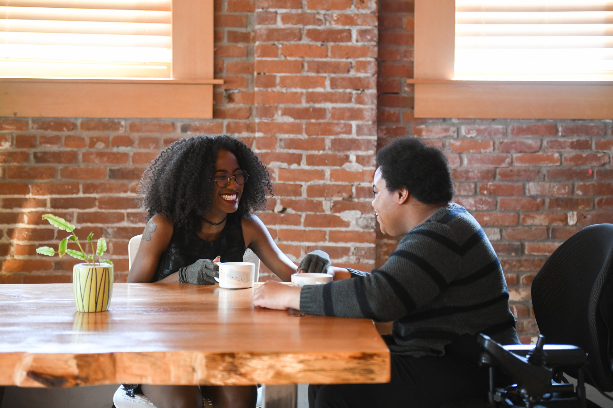 Two disabled Black people (a femme wearing compression gloves and a non-binary person in a power wheelchair that's partially in view) sit across each other and laugh while on a coffee date in a brick building. Photo from Disabled And Here: https://affecttheverb.com/disabledandhere/