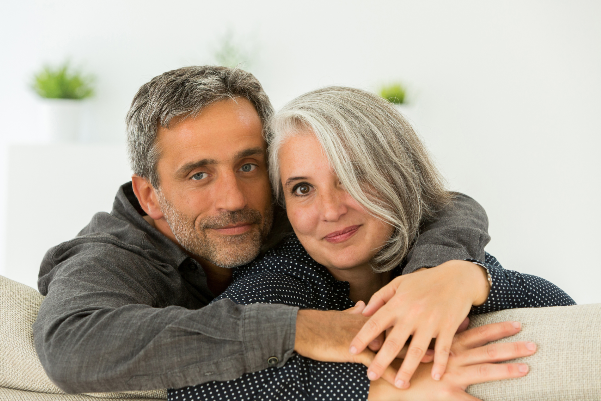 A middle-aged white woman with grey hair wearing a black, long-sleeved shirt with white polka dots and a middle-aged white man with grey hair wearing a grey, long-sleeved shirt, sitting next to each other and facing the camera while leaning against the back of a couch.