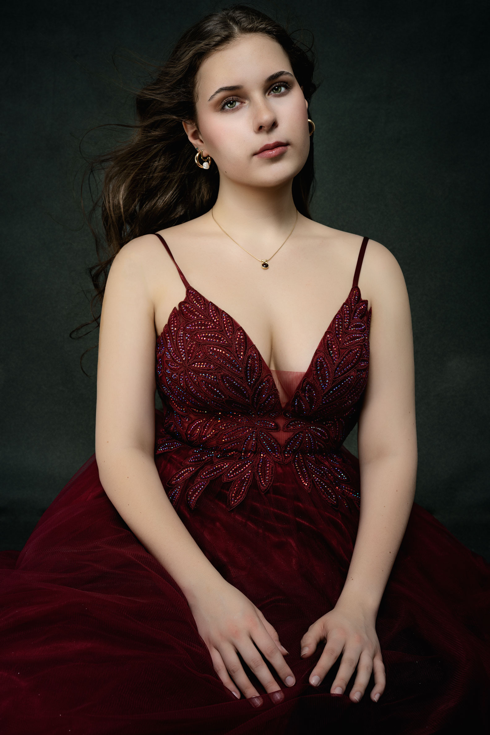 Editorial studio portrait of a woman wearing a burgundy dress seated against a dark background during a Portraits of Becoming session by Janice Smith Photography.