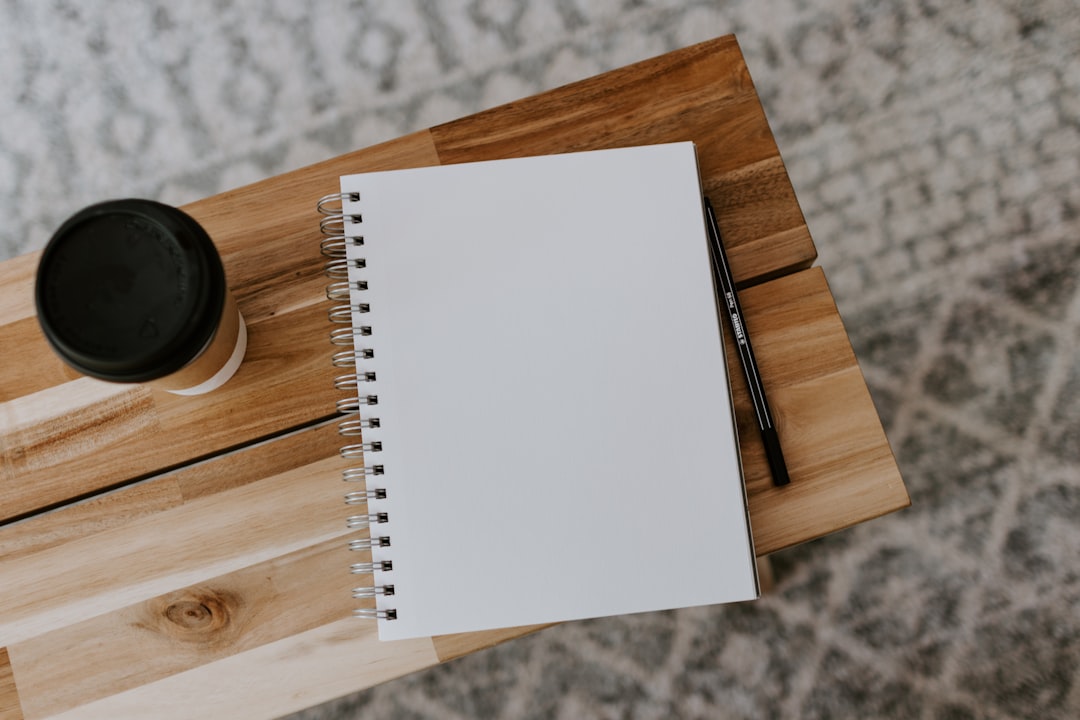 Overhead view of a blank spiral notebook, black pen and lidded coffee cup on a light wooden table over a patterned rug.
