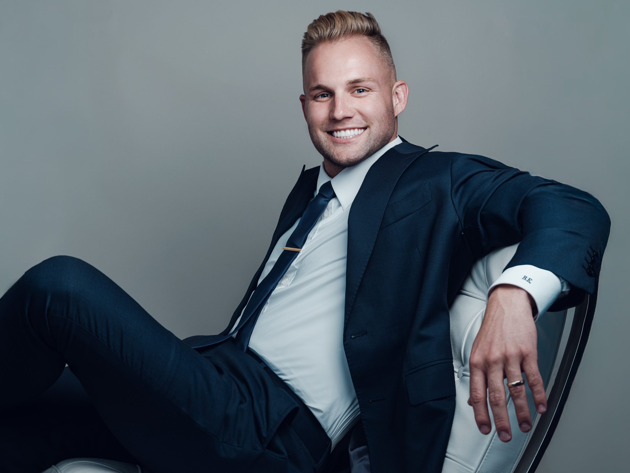 Blake Erickson in a navy suit, seated in a white chair, smiling
