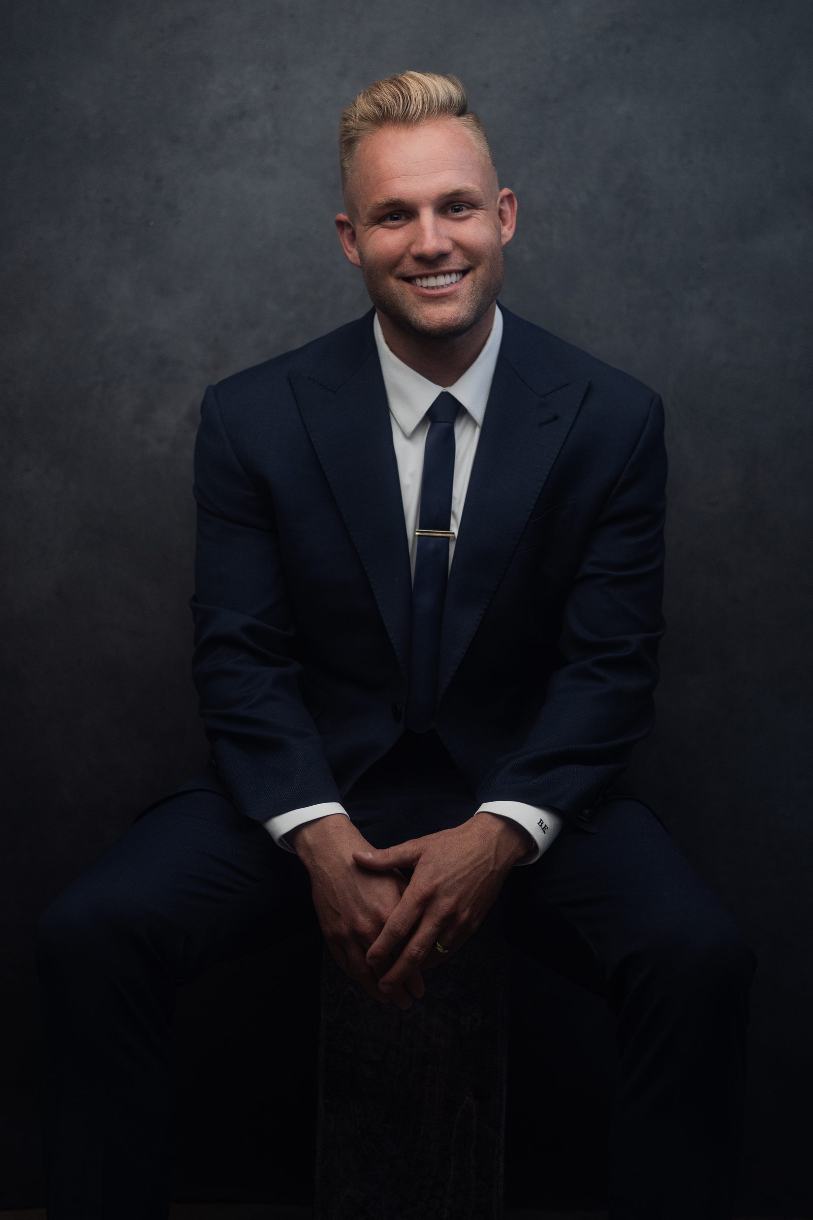 Blake Erickson in a navy suit, seated on a stool against a dark studio background, smiling