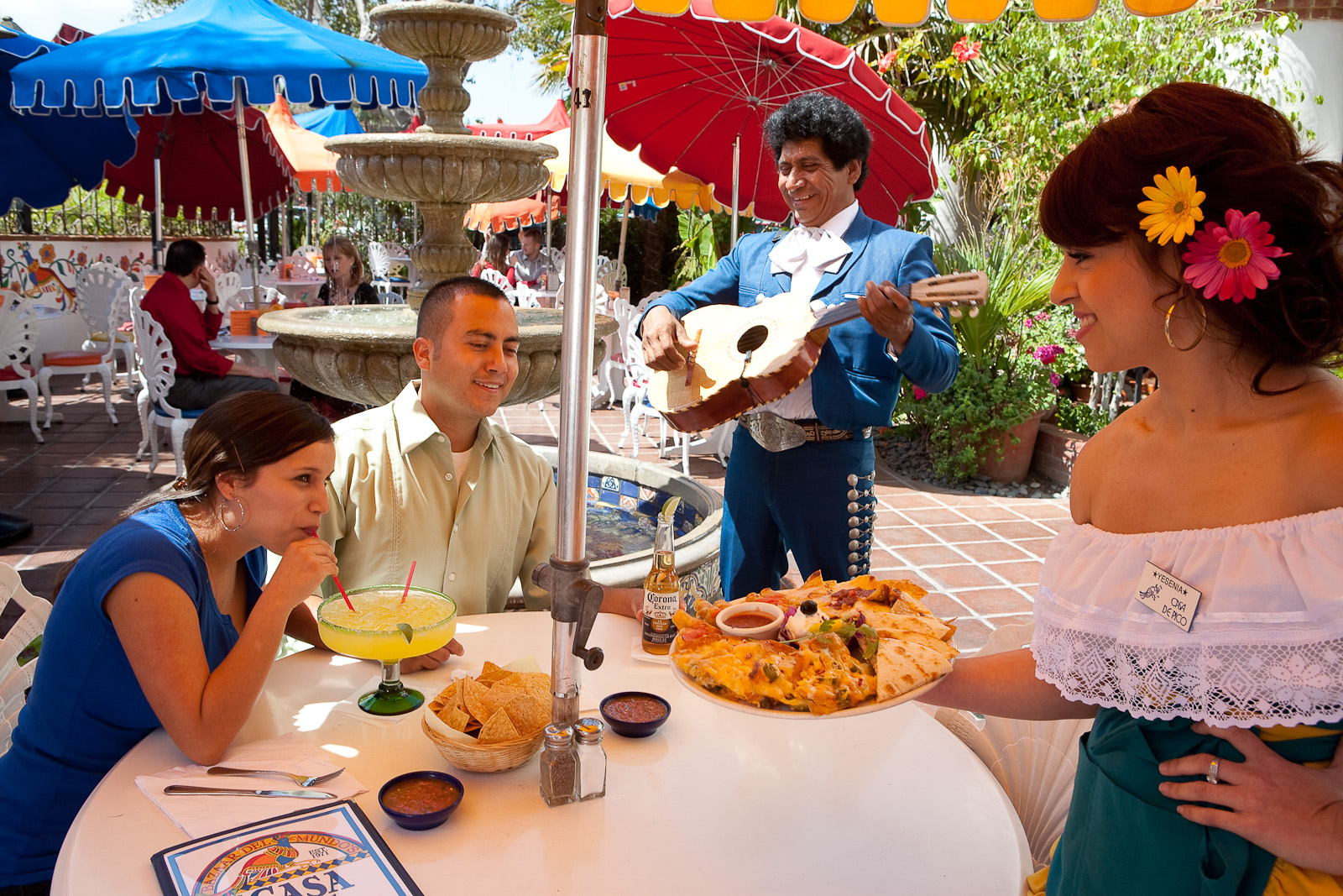 Lively Mexican restaurant patio at sunset with colorful umbrellas, guests enjoying sizzling fajitas and margaritas, string lights overhead, servers carrying large platters