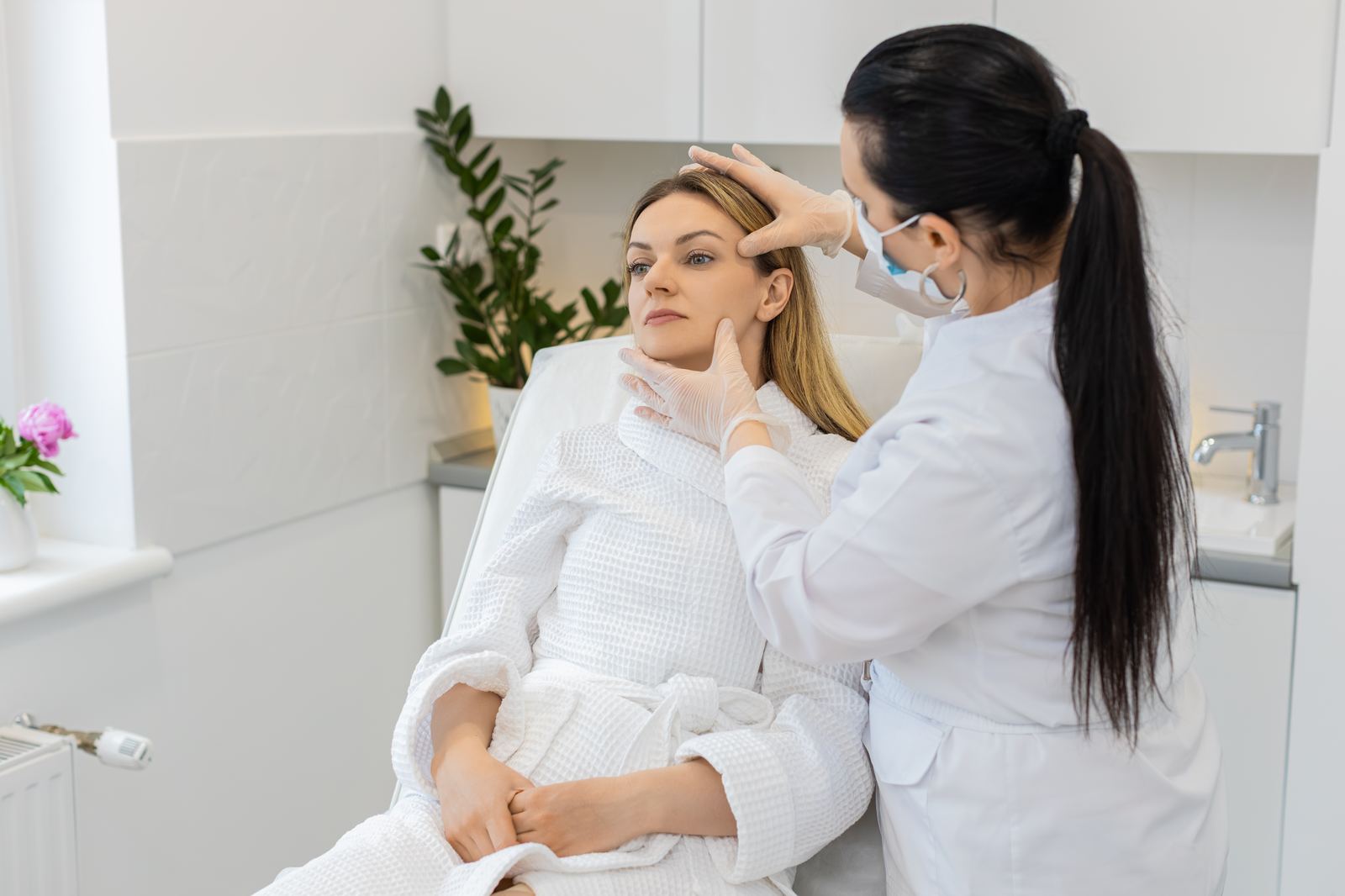 Cosmetóloga con guantes realizando una valoración facial a una clienta en una cabina luminosa