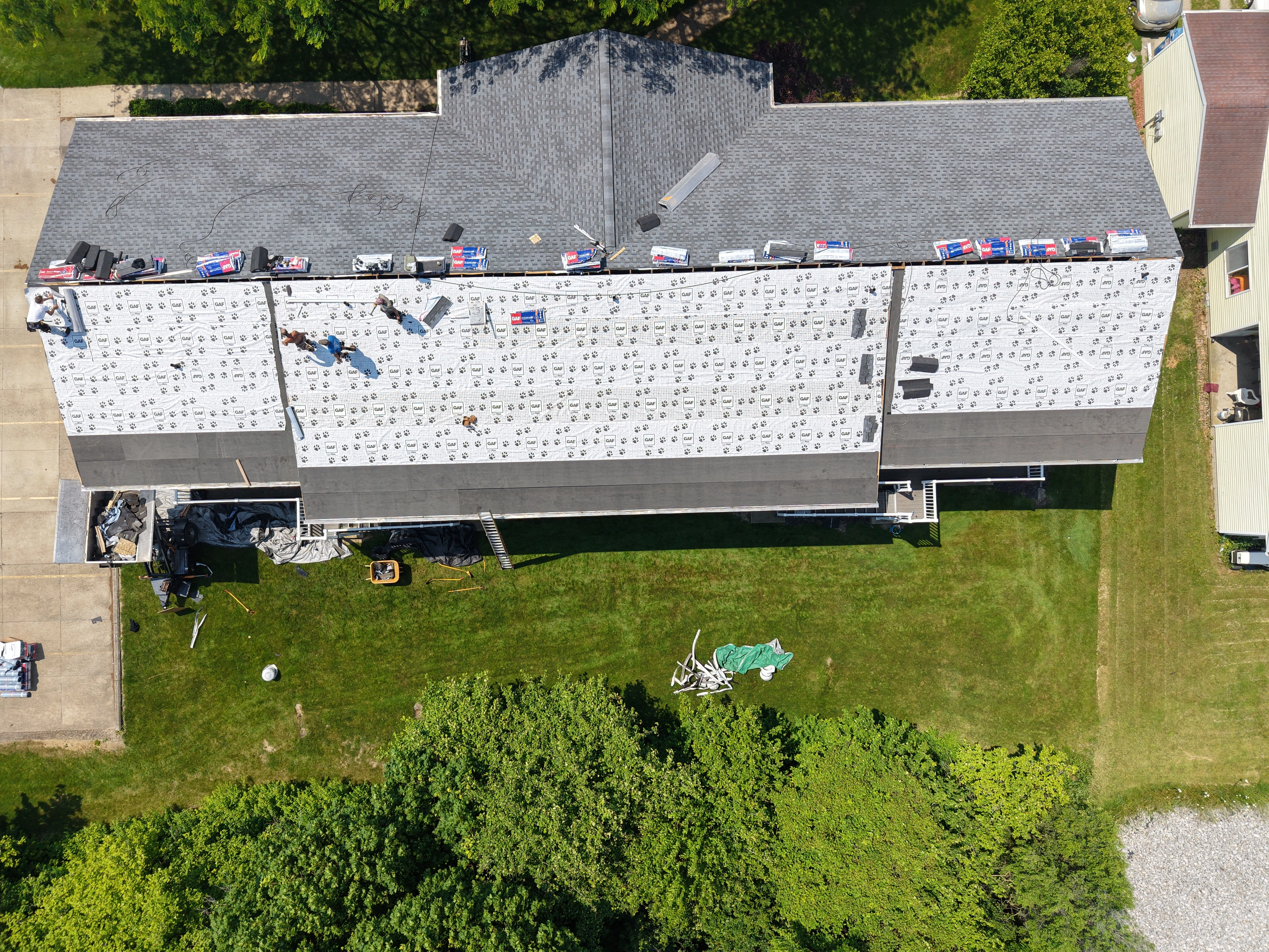Aerial view of a residential roofing project in progress, showing workers installing new shingles on a large home with white underlayment visible and roofing materials staged along the roof edge. - Weikles Roofing