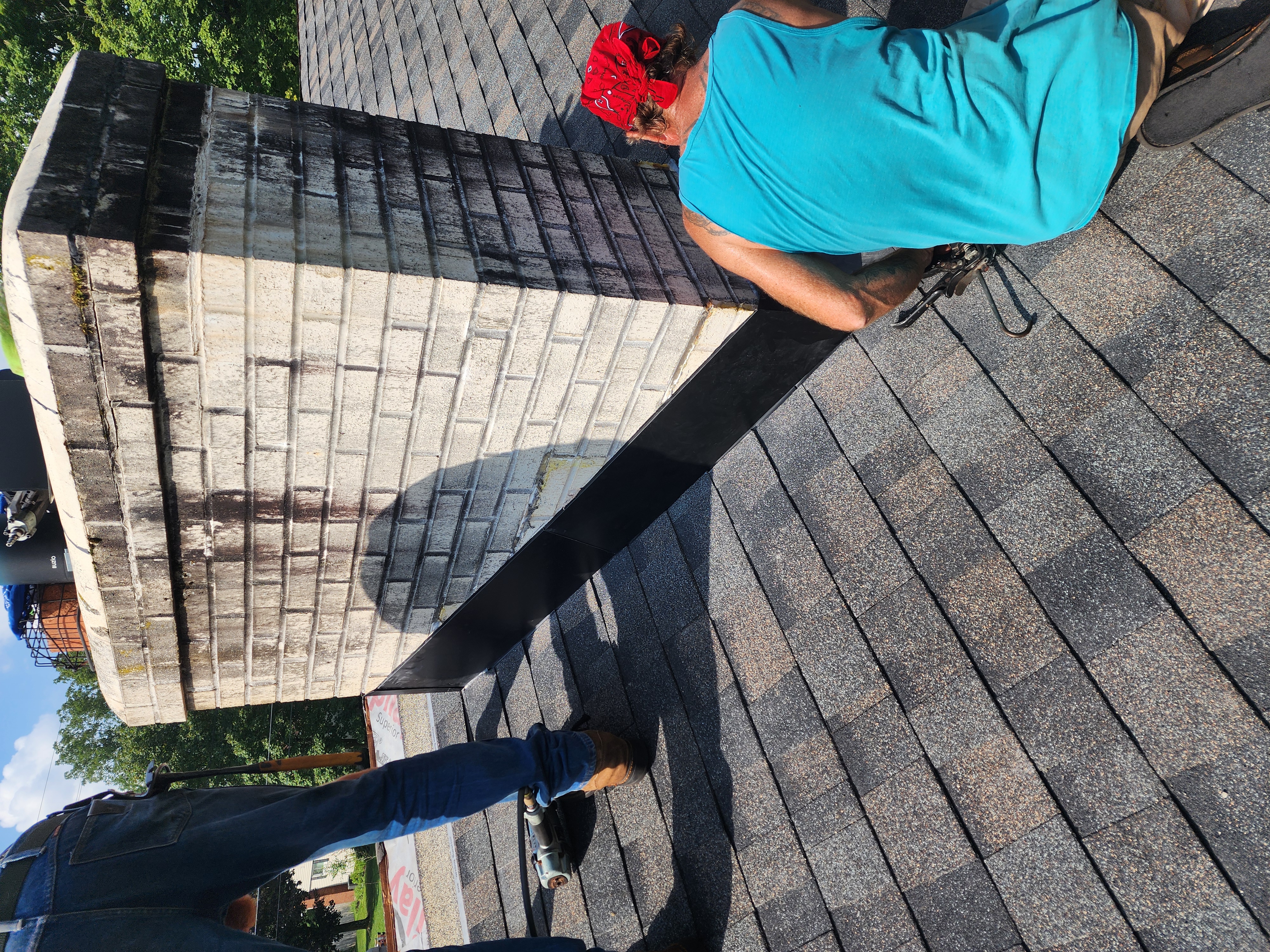 Two roofers are installing new metal flashing along a brick chimney on a residential roof with asphalt shingles. The work appears to be in progress, with one worker holding a piece of dark metal step flashing against the chimney during installation. - Weikles Roofing Two roofers are installing new metal flashing along a brick chimney on a residential roof with asphalt shingles. The work appears to be in progress, with one worker holding a piece of dark metal step flashing against the chimney during installation. - Weikles Roofing
