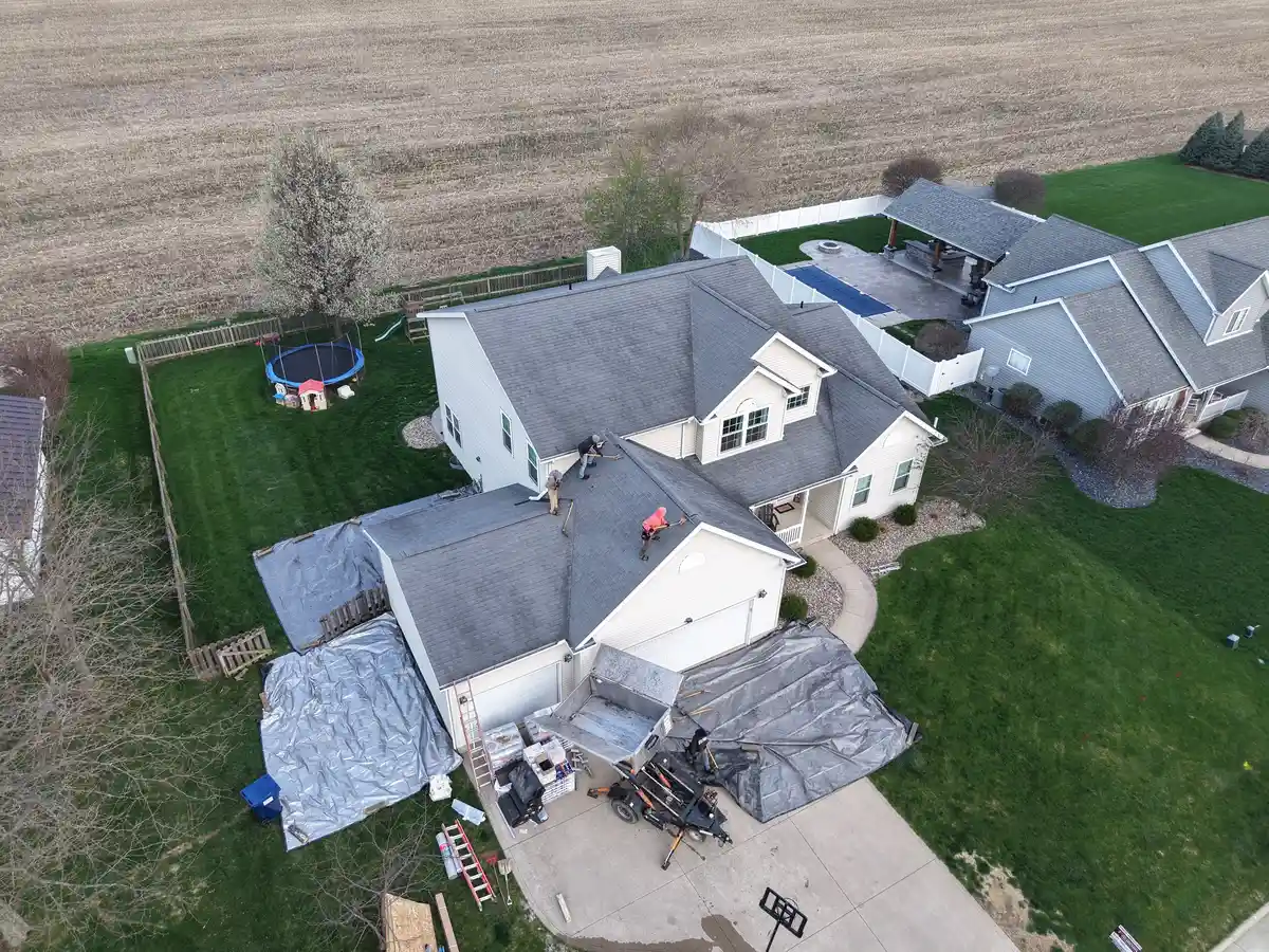Aerial view of a roofing crew removing shingles from a roof of a house on West Crest Drive in Shelby, Ohio, with debris and equipment on the driveway and tarps in the yard. - Weikles Roofing