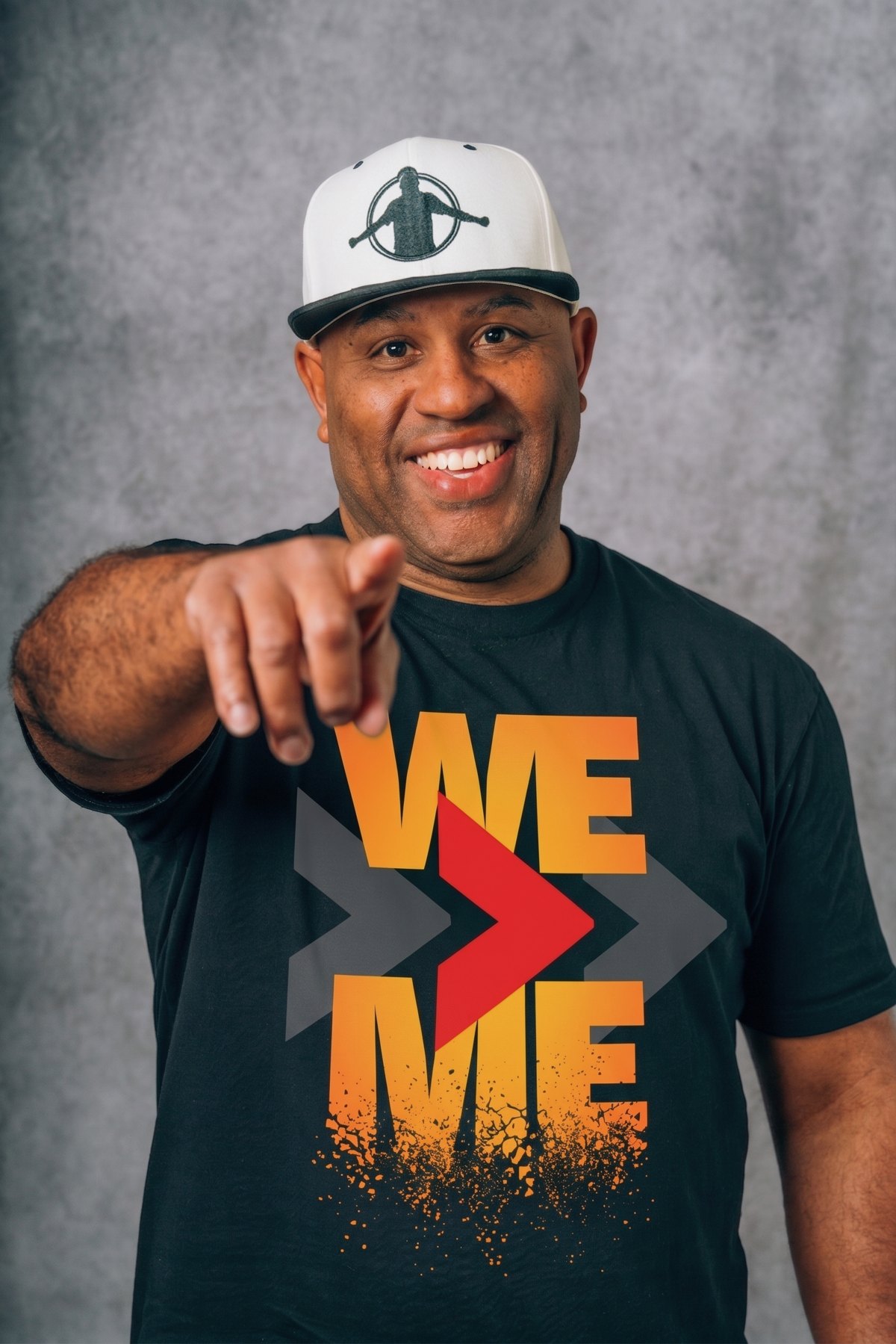 ET smiling and pointing at the camera in a We Is Greater Than Me shirt