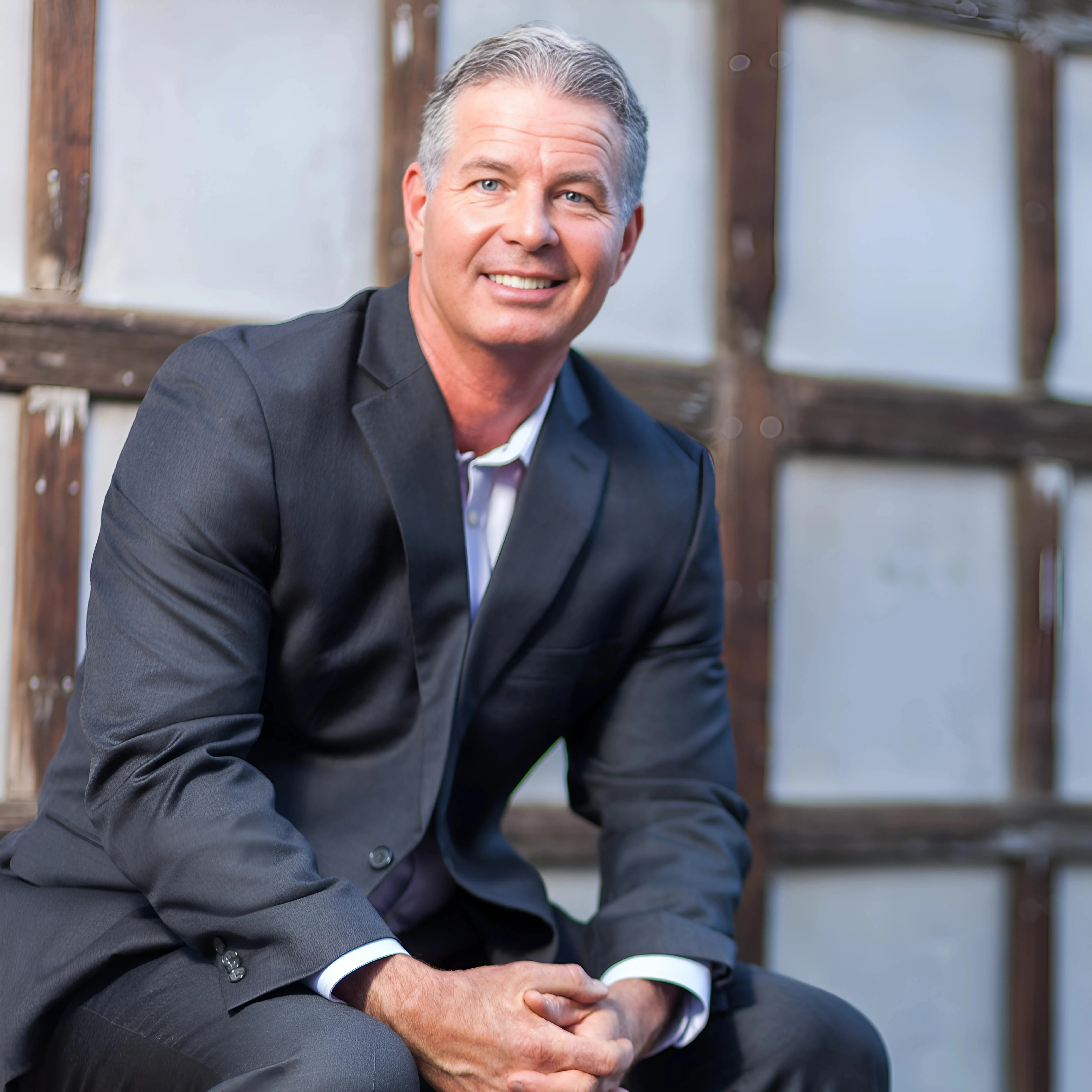 Professional man in a suit smiling confidently, seated against a rustic wooden backdrop, representing expertise in digital marketing and business growth.