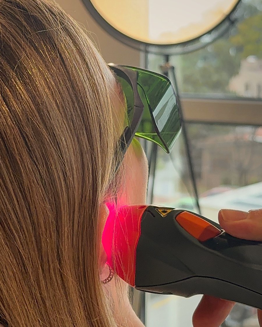 Paciente con anteojos de protección recibiendo una aplicación de láser terapéutico junto al oído, frente a una ventana