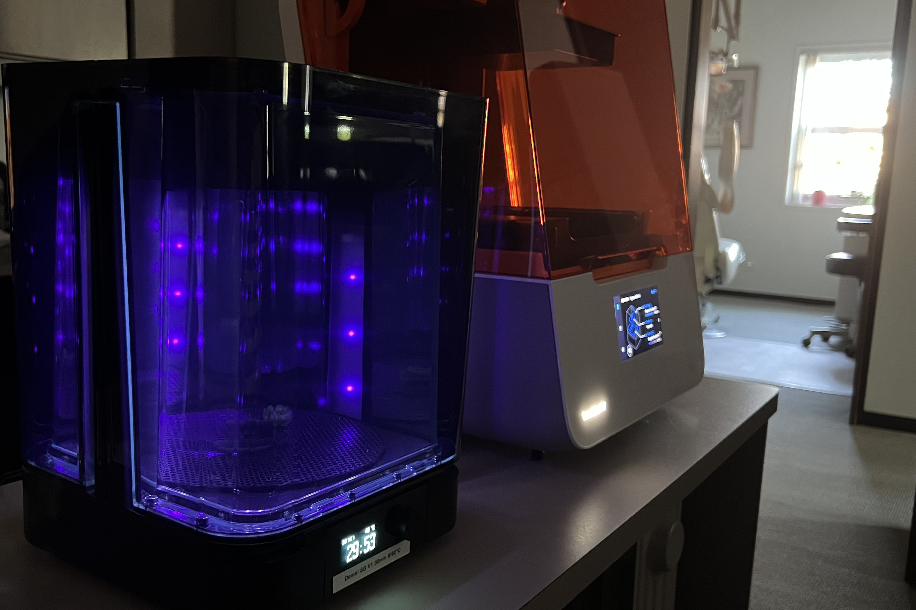 A picutre of a 3D printer and post cure machine sitting on a table post cure machine is illuminated and in use