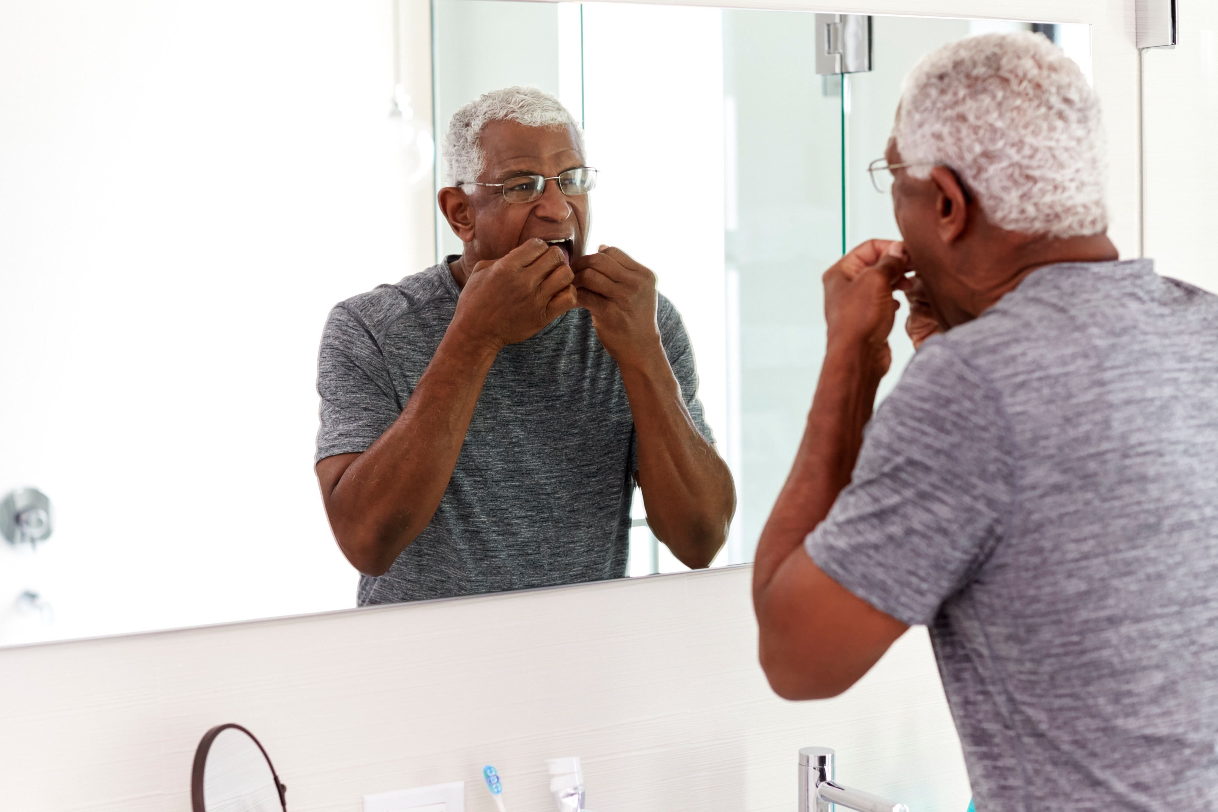 Old African American man with short white curly hair looking in the mirror flowing his teeth 