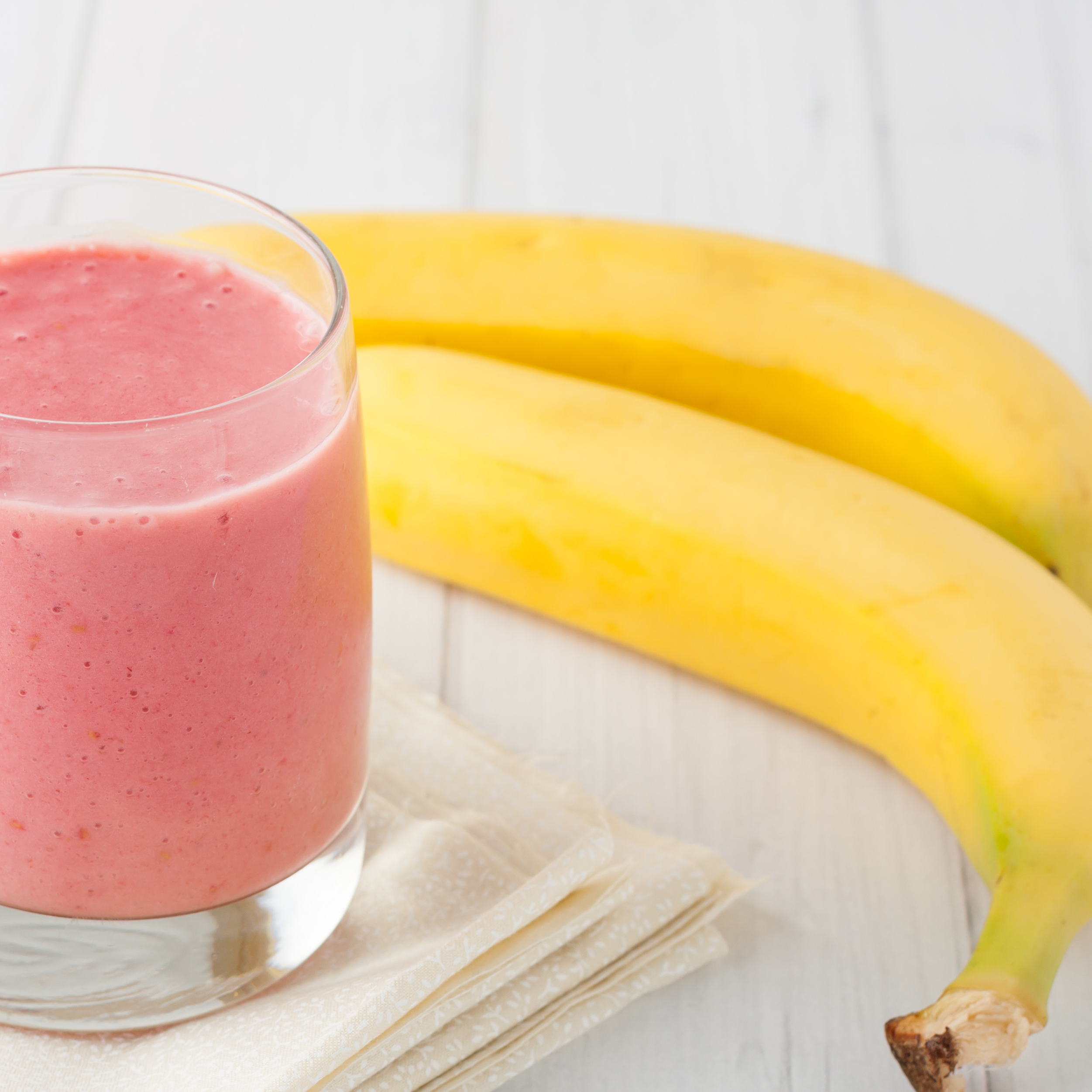 A photo of a no seed glass of strawberry banana smoothie next to two whole bananas and a white napkin, representing nutritious and soft food options for post-operative dental care.
