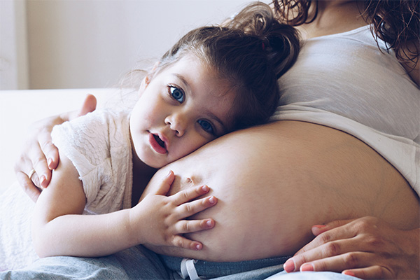 Pregnant Woman with her belly exposed and a white sports bar on with a young girl holding her ear and hand against the pregnant belly, looking directly into the camera. Depicting hormonal effects on gum health during pregnancy.