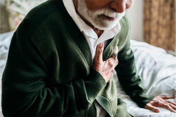 Elderly Indian man with a white beard clutching chest, wearing a white button-down with a dark green cardigan. Symbolizing the connection between heart disease and gum health.