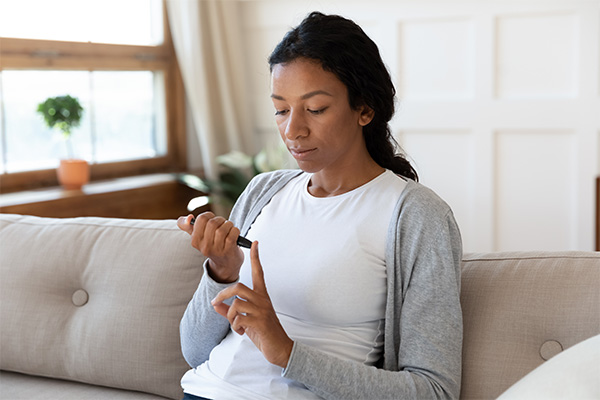 African American woman with hair pulled back in a ponytail sitting on a couch with a white shirt and grey cardigan checking glucose levels, linking diabetes management with periodontal care.