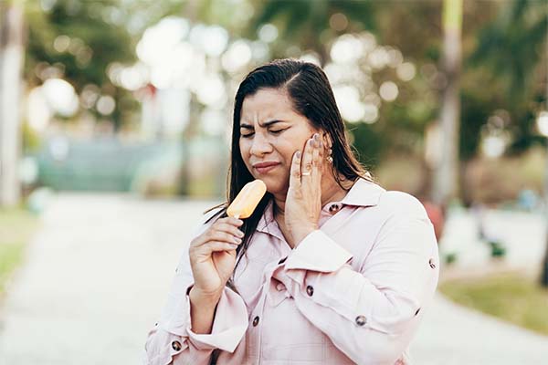 A Hispanic woman with black hair in a trenchcoat has a pained look on her face and is holding her jaw with one hand and a popsicle outdoors in a park, suggesting toothache or sensitivity from dental issues.