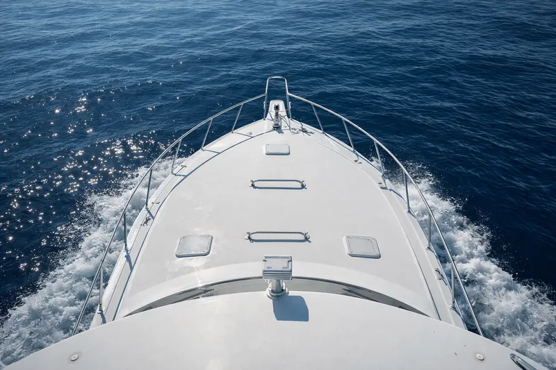Top-down view over the bow of the FishTriton sportfishing boat cruising on calm deep-blue ocean with wake foam and stainless bow rails visible.