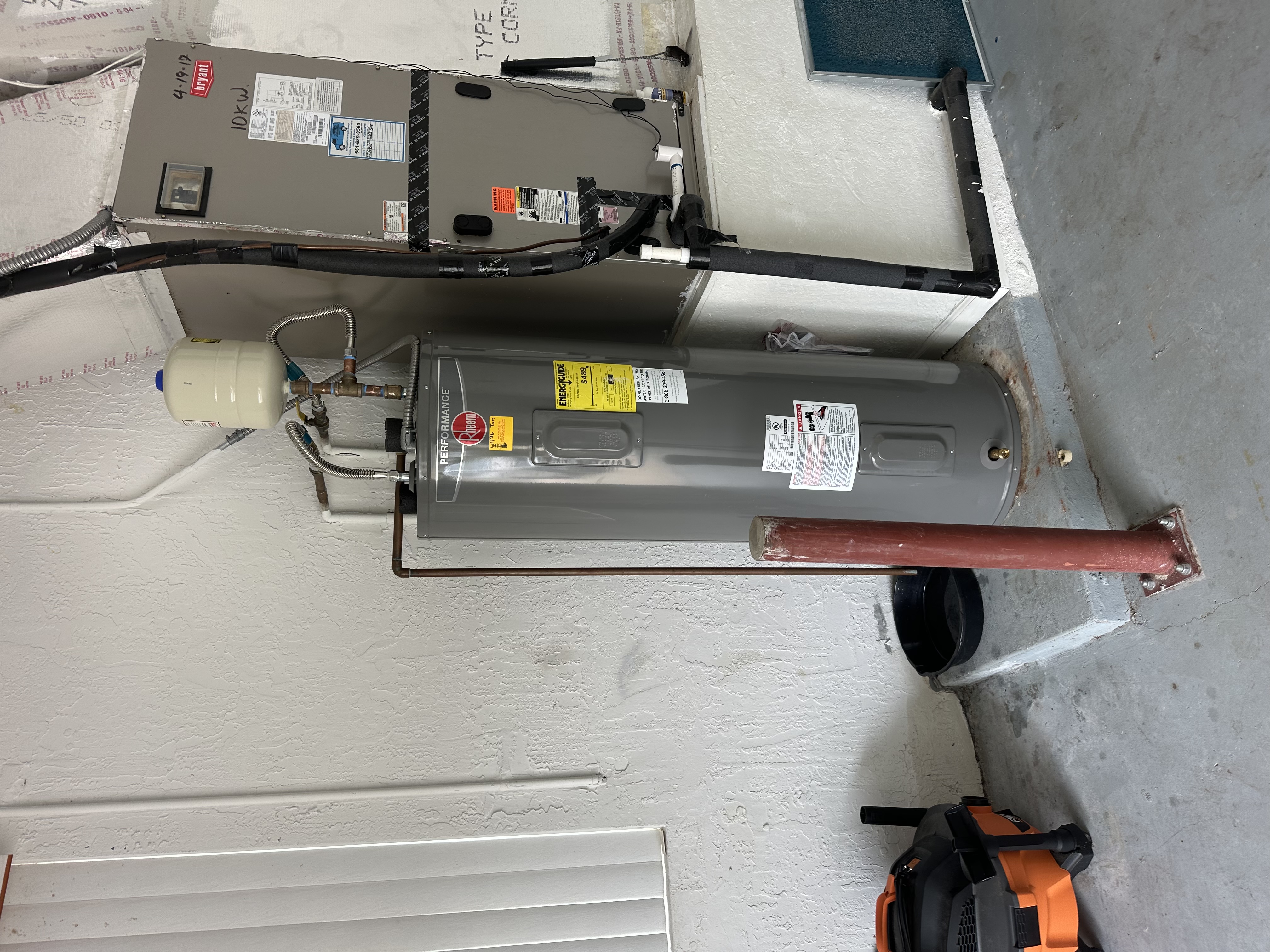 New Rheem Performance electric water heater installed in a garage beside the air handler, with an expansion tank mounted above it