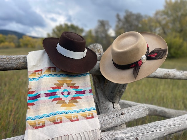 Brown felt hat and tan felt hat with a printed scarf, displayed with a woven blanket on a fence
