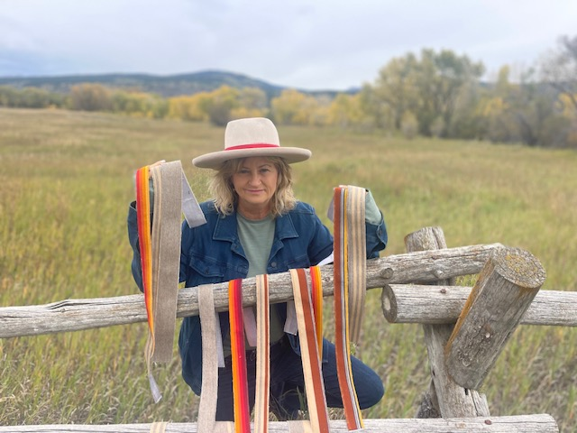 Woman in a cream felt hat with striped hatband ribbons on a fence, open field behind her