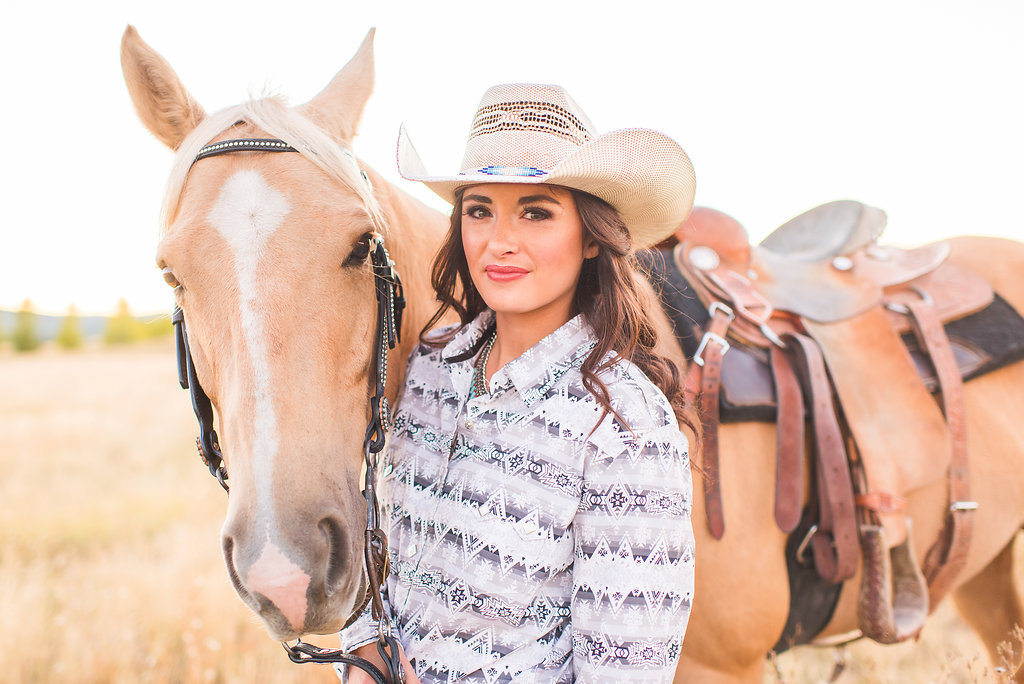Woman in a straw hat standing beside a saddled horse at golden hour