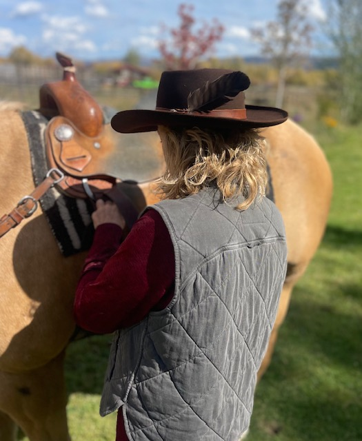 Woman in a feathered felt hat saddling a horse, seen from behind