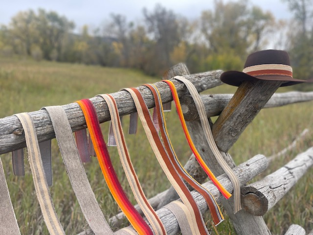 Rows of striped hatband ribbons on a wooden fence beside a dark felt hat