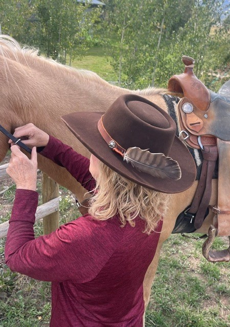 Woman in a feathered felt hat saddling a horse near an aspen grove
