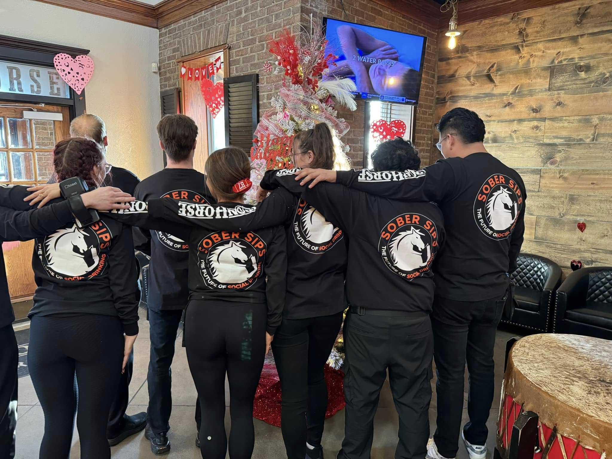 Group of people wearing matching Sober Sips shirts with the logo “The Future of Social Drinking,” standing together with arms around each other inside a bar. Tagline: Stay Sharp. Stay Social. Stay You.