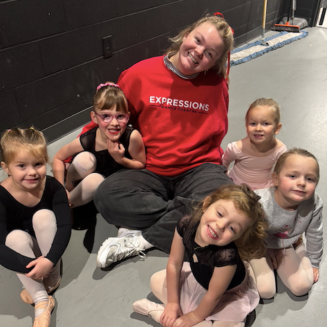 A dance teacher in a red “Expressions Dance Theatre” sweatshirt sits on the floor with five young girls in ballet clothes, all smiling at the camera.