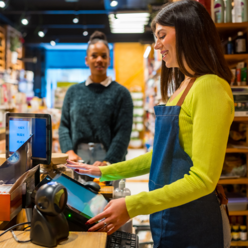 Smiling retail worker in a blue apron taps a touchscreen POS terminal at the checkout while a customer waits in the background.
