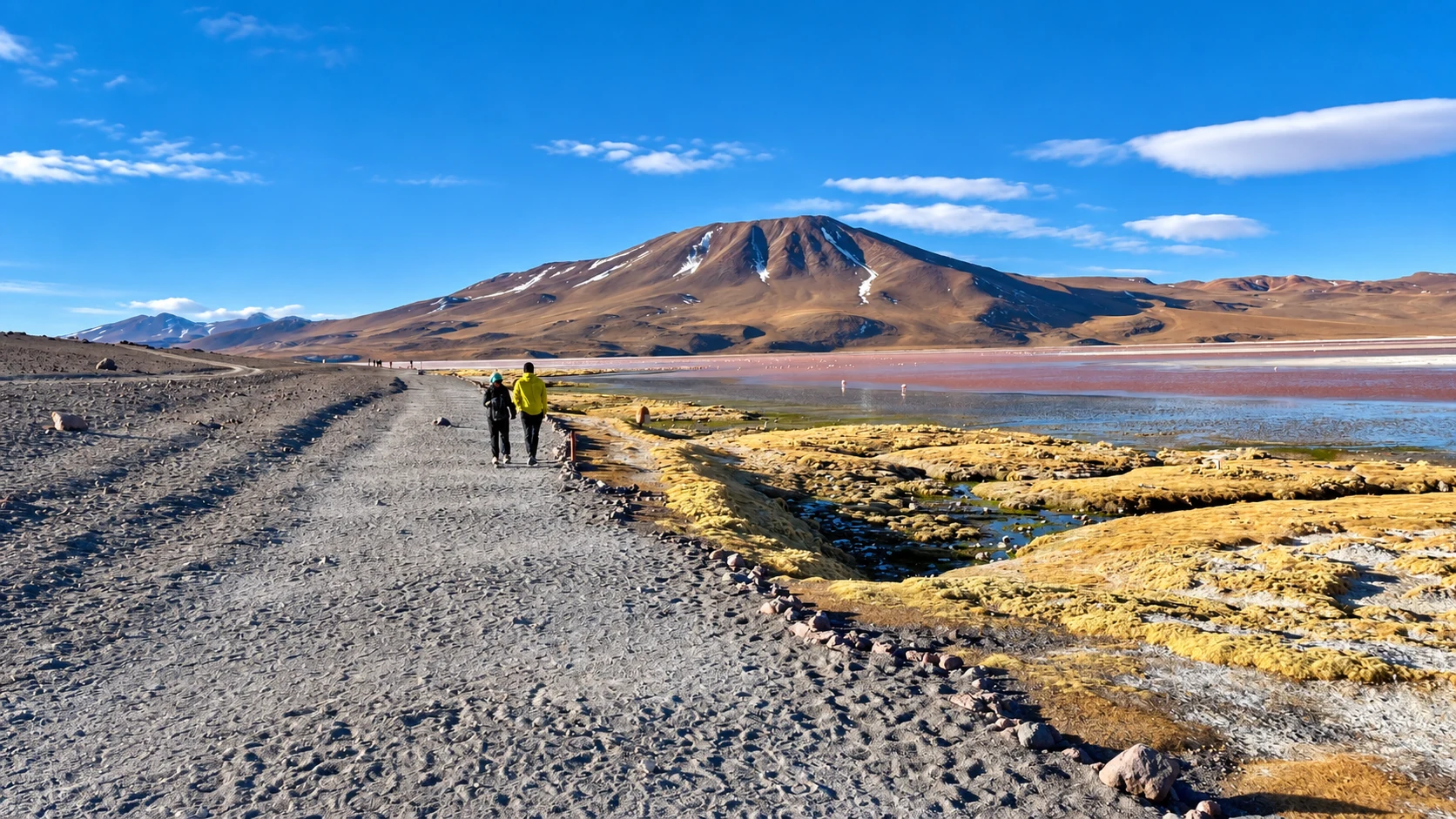 Viajeros caminando junto a una laguna del Altiplano con un volcán al fondo