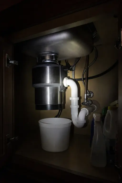 Garbage disposal and PVC drain trap beneath a kitchen sink with a bucket below in Southport, NC
