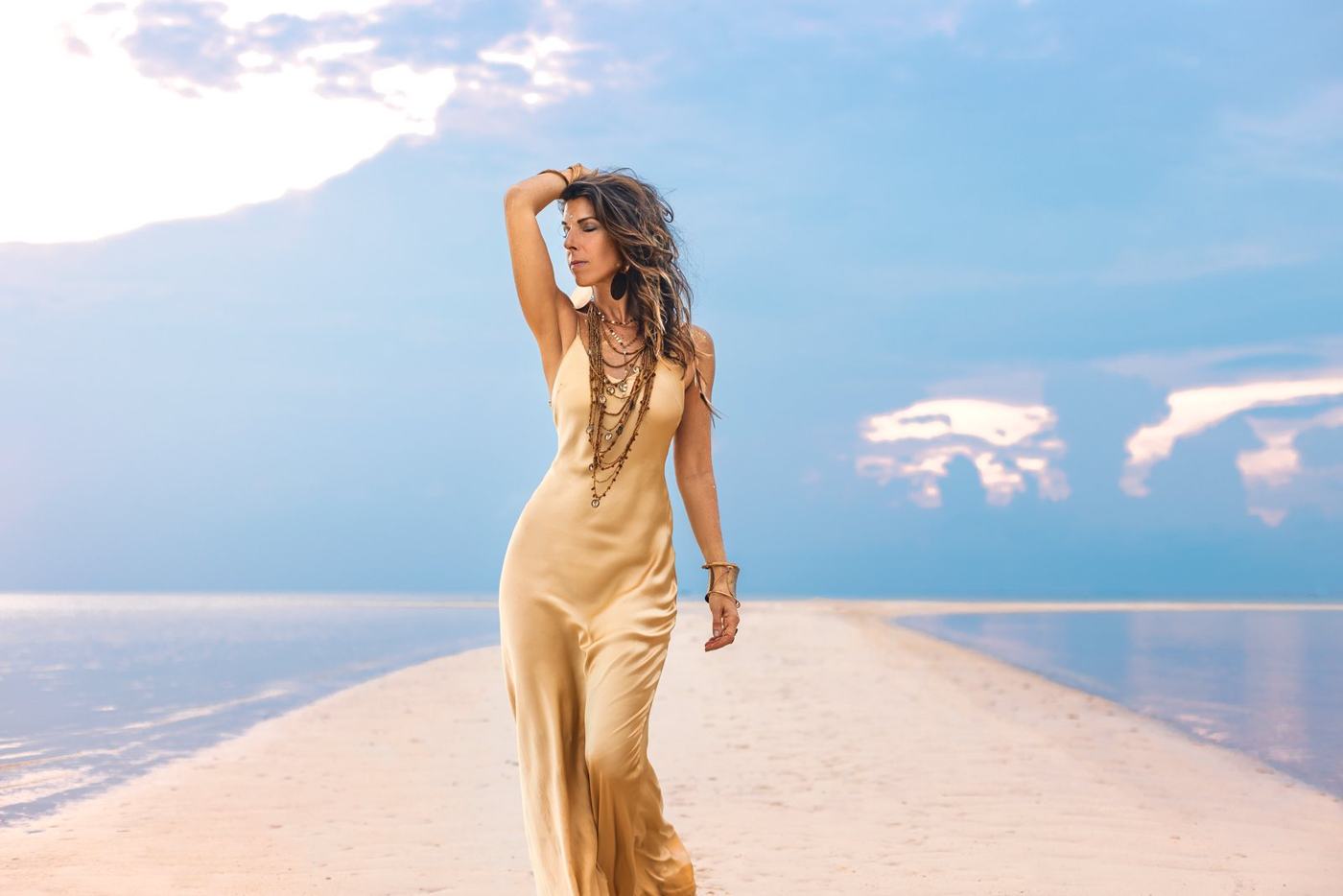 Nicole in a gold silk dress walking the sandbar, one hand in her hair, looking to the side