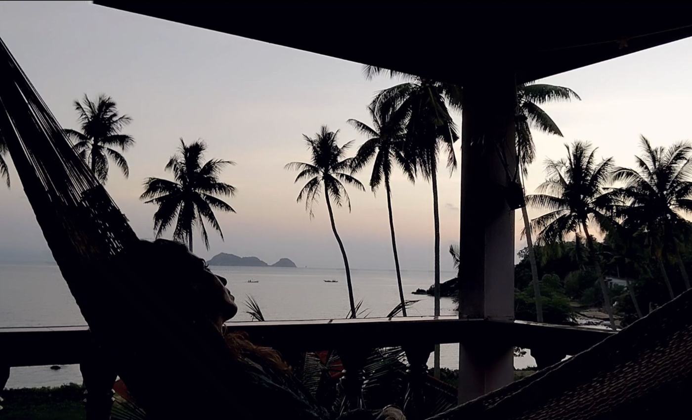 Nicole in silhouette resting in a rope hammock on a veranda at dusk, palms, the sea and a small island beyond under a lavender sky