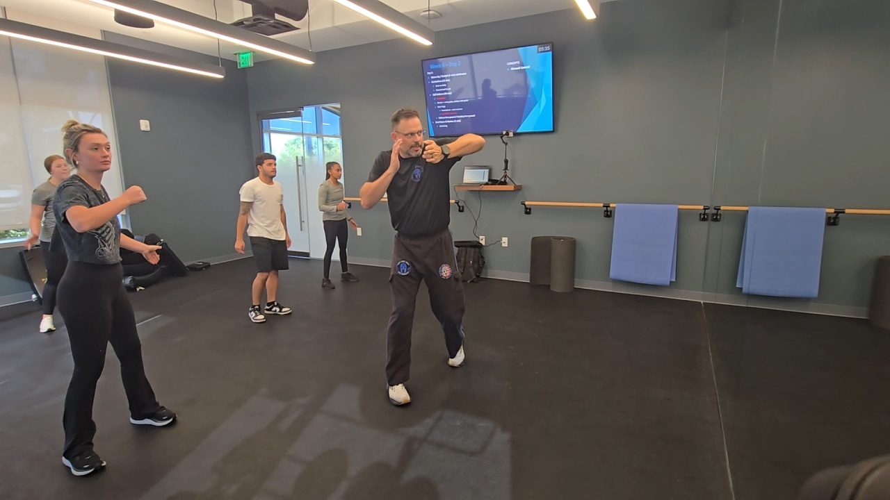 Jason Mertin mid-instruction, demonstrating a defensive block while students train behind him in the studio
