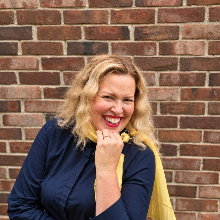 Mayra Sanchez laughing at a brick wall, in navy with a gold scarf