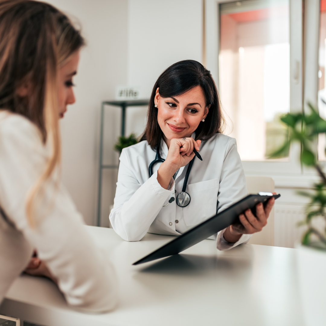 Female doctor smiling while discussing results with a patient, holding a clipboard during a consultation in a bright office.