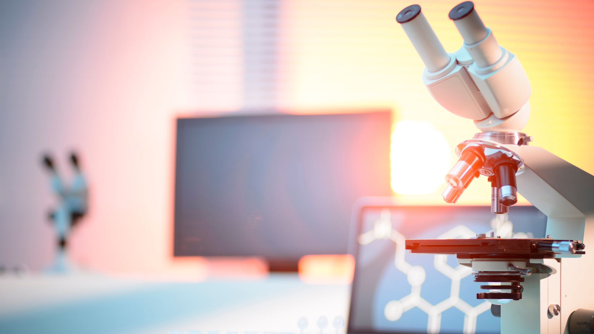 Microscope on a lab bench with test tubes and computer screens in a brightly lit modern laboratory.