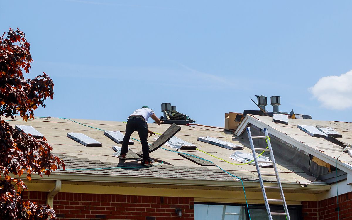 A roof replacement in progress on a brick home, the old shingles stripped back to bare deck and a ladder up to the eaves