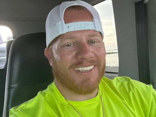 Smiling man wearing a white cap and neon yellow shirt while sitting inside a vehicle.